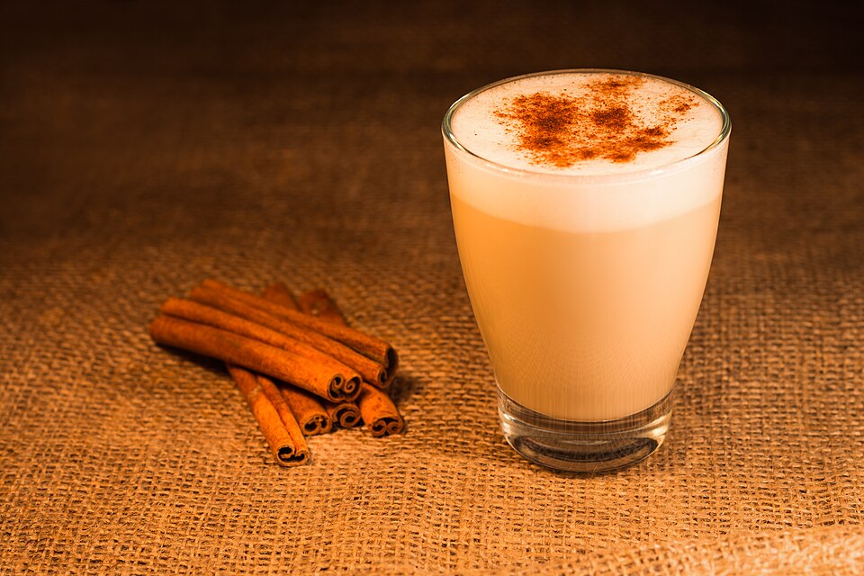 Latte con espuma y canela espolvoreada en vaso de vidrio, junto a rajas de canela